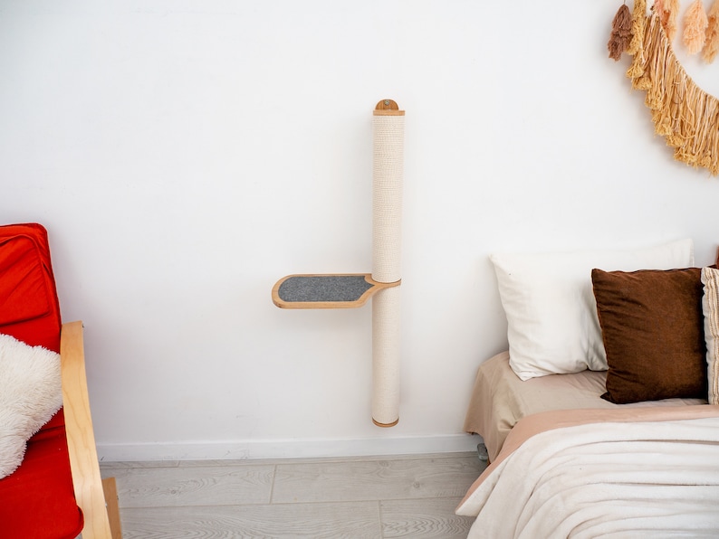 Pode incluir: Um arranhador de parede para gatos com uma plataforma. O poste &eacute; coberto com corda de sisal e a plataforma &eacute; coberta com tecido cinza. O poste &eacute; montado em uma parede branca e h&aacute; uma cama com almofadas no fundo.