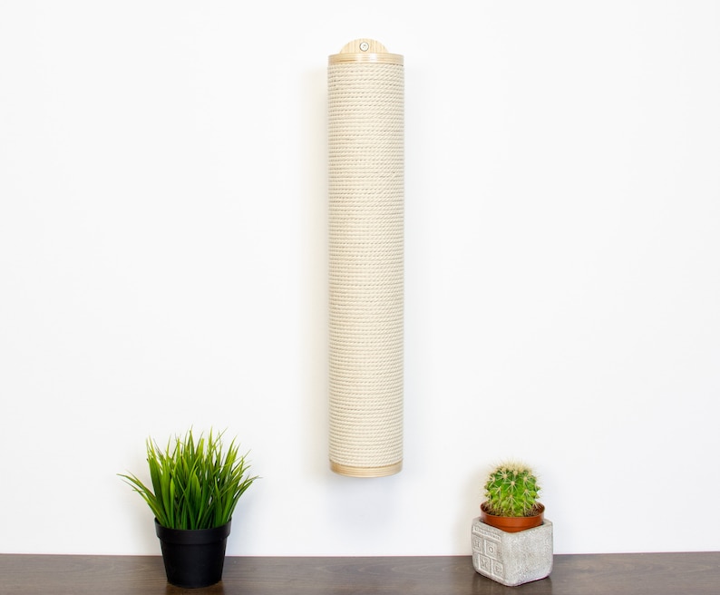 May include: A beige sisal scratching post for cats, mounted on a white wall. The post is made of natural sisal rope and has a wooden top. There are two potted plants on a wooden surface below the post.