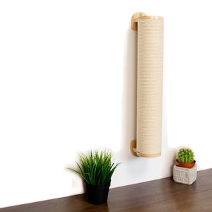May include: A beige scratching post for cats is mounted on a white wall. The post is made of natural sisal rope and has a wooden frame. A small potted plant sits on a wooden surface below the post.