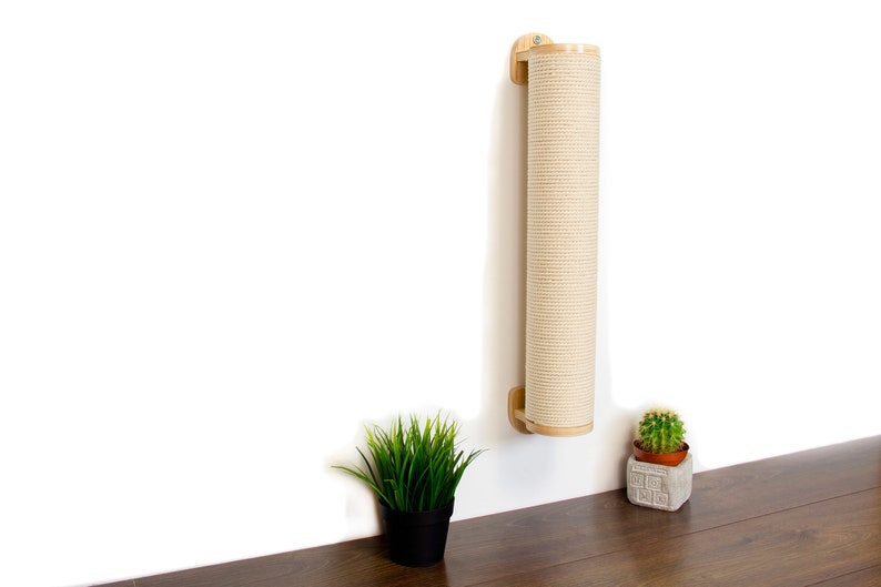 May include: A beige scratching post for cats is mounted on a white wall. The post is made of natural sisal rope and has a wooden frame. A small potted plant sits on a wooden surface below the post.