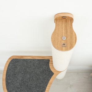 Pode incluir: Um arranhador para gatos branco e cinza com uma base de madeira e uma plataforma de feltro cinza. O poste &eacute; fixado a uma parede com um suporte de madeira. O suporte tem uma pequena etiqueta de madeira com o texto "Htt Hakata" nela.