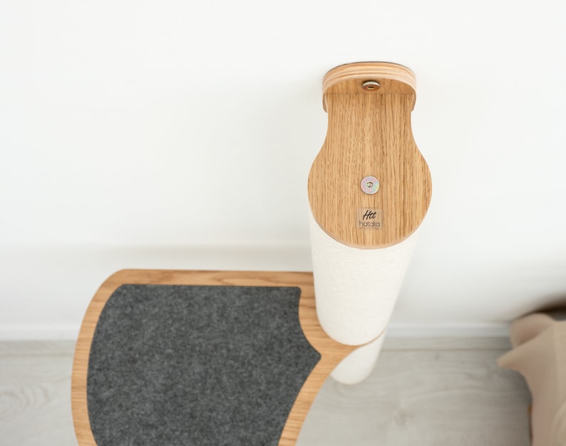 Pode incluir: Um arranhador para gatos branco e cinza com uma base de madeira e uma plataforma de feltro cinza. O poste &eacute; fixado a uma parede com um suporte de madeira. O suporte tem uma pequena etiqueta de madeira com o texto "Htt Hakata" nela.