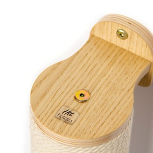 May include: A wooden cat scratching post with a natural wood finish and a beige rope covering. The post is mounted to a wall with two screws. The post has a small label with the text "Htt hatata" on the side.