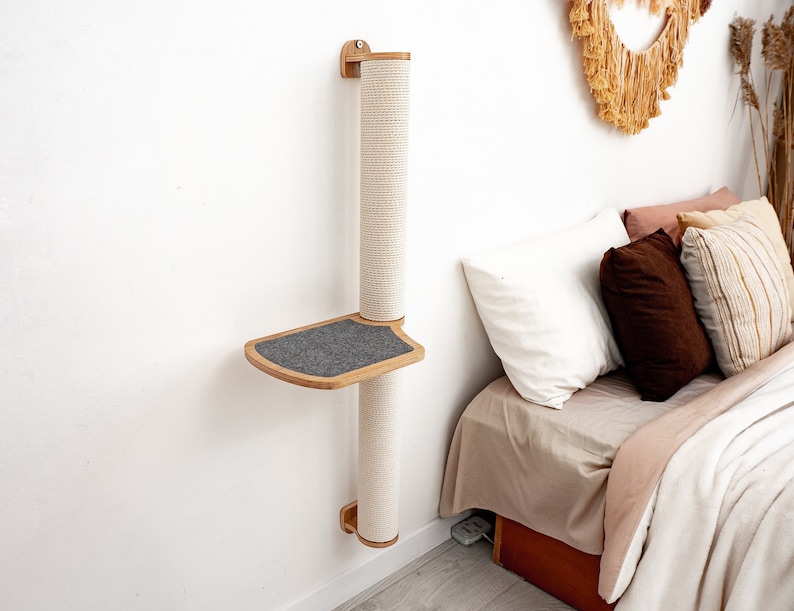 Pode incluir: Um arranhador de gato de parede com uma plataforma e corda de sisal. O poste &eacute; feito de madeira natural e tem uma plataforma cinza. O poste est&aacute; montado em uma parede branca ao lado de uma cama com almofadas.