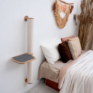 Pode incluir: Uma &aacute;rvore de gato de parede com um poste para arranhar e uma plataforma. A &aacute;rvore de gato &eacute; feita de madeira e tem uma plataforma cinza. A &aacute;rvore de gato est&aacute; montada em uma parede branca ao lado de uma cama com roupa de cama e travesseiros brancos.