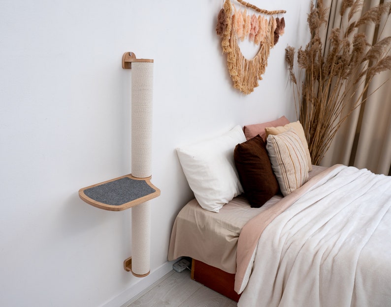Pode incluir: Uma &aacute;rvore de gato de parede com um poste para arranhar e uma plataforma. A &aacute;rvore de gato &eacute; feita de madeira e tem uma plataforma cinza. A &aacute;rvore de gato est&aacute; montada em uma parede branca ao lado de uma cama com roupa de cama e travesseiros brancos.