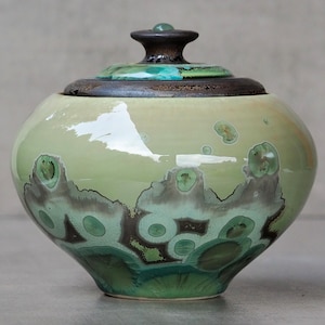 Op de afbeelding: Een ronde keramische pot met deksel, voorzien van een glanzend lichtgroen glazuur met donkergroene en bruine kristallijne patronen. Het deksel heeft een donkerbruine, getextureerde rand en een kleine knop als handvat.