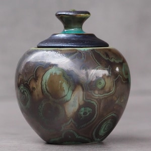 May include: A ceramic jar with a lid. The jar is a rounded shape with a dark brown and green mottled pattern. The lid is dark blue with a green knob. The jar has a glossy finish.