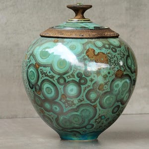 Op de afbeelding: Een ronde keramische pot met deksel en een uniek malachietpatroon in tinten van teal, bruin en groen. De pot heeft een bronskleurige rand en een kleine, bruine handgreep op het deksel. De basis van de pot heeft een teal-kleurige band.
