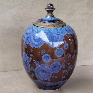 May include: A ceramic jar with a lid featuring a crystalline glaze. The body is a deep brown color with blue, circular, starburst-like crystal patterns. The lid has a matching blue and brown glaze with a small, dark, spherical finial on top.