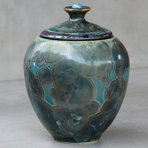Op de afbeelding: Een handgemaakte keramische pot met een deksel. De pot heeft een afgeronde vorm, met een turquoise en blauwe kristallijne glazuur. Het deksel heeft een kleine knop en een donkerdere turquoise rand. Het is een decoratief item.