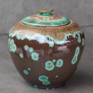 Op de afbeelding: Een bruine keramische pot met deksel, versierd met turquoise en beige accenten. Het deksel is groen met een gouden rand. De pot heeft een ronde vorm en een glanzende afwerking. Waarschijnlijk voor opslag of decoratie. Hoogte ongeveer 15 cm.