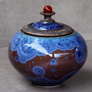 Può includere: Un barattolo in ceramica rotondo con coperchio, caratterizzato da una smaltatura di base marrone scuro con vivaci motivi cristallini blu che sbocciano sulla superficie. Il coperchio è sormontato da una piccola impugnatura sferica rossa, liscia e lucida.