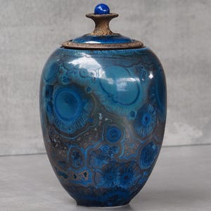 Op de afbeelding: Een keramische pot met deksel voorzien van een kristallijn glazuur in tinten diepblauw, turkoois en donkerbruin. Het oppervlak vertoont overal cirkelvormige, stervormige patronen. Een kleine, massieve blauwe bol zit bovenop de handgreep van het deksel.