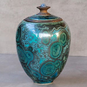 Peut inclure: Un pot en céramique avec couvercle présentant une finition brillante et un motif inspiré de la malachite dans des tons de bleu sarcelle, turquoise et brun foncé. Le récipient a un corps arrondi et un petit bouton sphérique sur le couvercle.