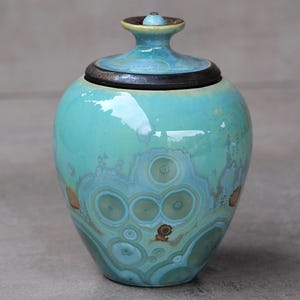 May include: A turquoise ceramic jar with a lid. The jar has a round body and a lid with a small knob. The jar's surface features a unique pattern of concentric circles in shades of blue and green. The lid is dark brown.