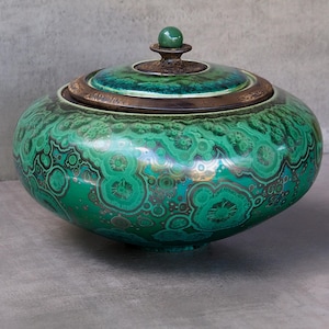 Op de afbeelding: Een ronde keramische pot met deksel, voorzien van een levendig groen en blauwgroen kristallijn glazuur dat het uiterlijk van malachiet nabootst. Het deksel heeft een donkerbronskleurige rand en een kleine, gladde, bolvormige groene knop.