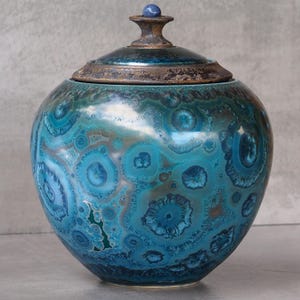Op de afbeelding: Een ronde keramische pot met deksel, voorzien van een kristallijn glazuur in tinten diepblauw, turkoois en bruin. Het oppervlak vertoont ingewikkelde, stervormige kristalpatronen, bekroond met een donker, getextureerd deksel en een kleine blauwe knop.