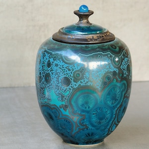 Op de afbeelding: Een keramische pot met deksel voorzien van een kristallijn glazuur in tinten teal, turquoise en donkerbruin. Het oppervlak vertoont overal cirkelvormige, stervormige patronen. Bovenop de dekselknop zit een kleine, gladde, lichtblauwe bol.
