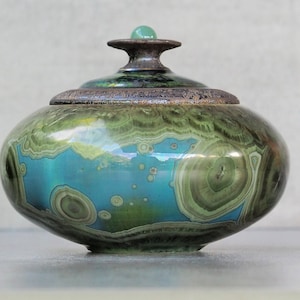 Op de afbeelding: Een ronde keramische pot met deksel, voorzien van een glanzende afwerking met wervelende patronen in tinten diep bosgroen, olijfgroen en levendig turkoois. Het deksel heeft een donkere, getextureerde rand en een kleine, gladde, lichtgroene knop.