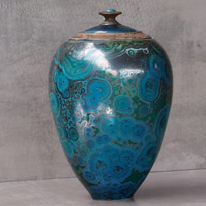 Op de afbeelding: Een keramische urn met deksel, voorzien van een glanzende afwerking in tinten blauw, turkoois en petrol. Het oppervlak vertoont complexe, cirkelvormige en organische patronen die doen denken aan malachiet, met een donkerbruine, getextureerde rand.