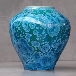 Puede incluir: Un jarrón decorativo de cerámica con una superficie texturizada única. El jarrón presenta una mezcla de tonos turquesa y azul, con un patrón de diseños circulares. El jarrón tiene un cuerpo redondeado y un borde ligeramente ensanchado.