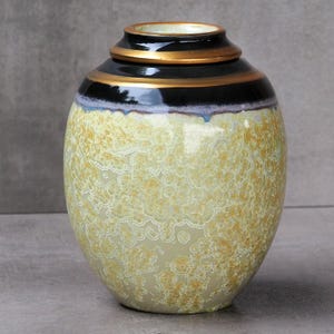 Op de afbeelding: Een decoratieve keramische vaas met een afgeronde body en een smalle hals. De vaas heeft een getextureerd, geel en crèmekleurig oppervlak, een zwarte band en gouden rand rond de bovenkant. Een decoratief woonaccessoire.