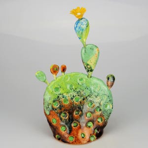 Peut inclure: Une sculpture de cactus en céramique aux couleurs vives. La base est de forme arrondie avec un glaçage moucheté vert et marron, comportant plusieurs ouvertures circulaires. Le cactus a plusieurs segments verts et bleus, surmontés d'une fleur jaune.