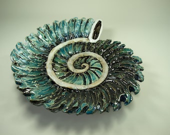 Puede incluir: Un plato de cerámica con forma de concha de nautilus con un esmalte azul y verde. La concha está detallada con un borde blanco y un patrón en espiral.