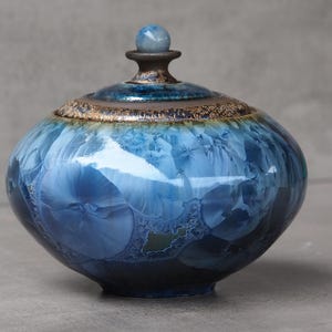 May include: A round ceramic jar with a lid featuring a crystalline glaze in shades of deep blue and light blue. The surface displays distinct starburst patterns. The lid has a dark brown rim with a textured finish and a small, smooth blue sphere knob.