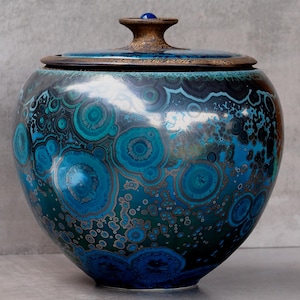 May include: A round ceramic jar with a lid featuring a crystalline glaze in shades of deep blue, turquoise, and black. The surface displays circular patterns resembling malachite or agate. A small blue knob sits atop the textured brown lid.