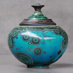 Puede incluir: Un tarro de cerámica redondo con tapa, con un esmalte vibrante en tonos turquesa y verde azulado. La superficie está decorada con patrones circulares de color verde oscuro y negro que recuerdan a la malaquita. La tapa tiene un borde oscuro.