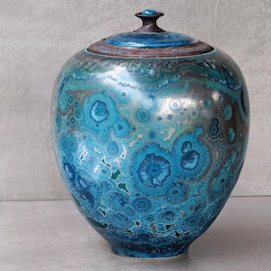 May include: A ceramic jar with a lid featuring a crystalline glaze in shades of vibrant turquoise, deep blue, and metallic bronze. The surface displays intricate, star-like crystal formations scattered across the rounded body and matching lid.