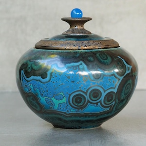 Może przedstawiać: Okrągły ceramiczny pojemnik z pokrywką, ozdobiony żywym szkliwem z wirującymi wzorami w kolorach turkusowym, głębokiego błękitu i czerni. Pokrywka ma teksturowaną krawędź w kolorze brązu oraz mały, gładki, jasnoniebieski kulisty uchwyt.