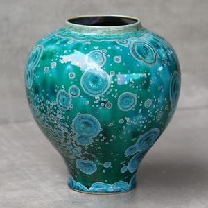 May include: A handcrafted ceramic vase with a rounded body and a narrow neck. The vase features a vibrant teal glaze with crystalline patterns in shades of blue and white. The vase is approximately 10 inches tall.