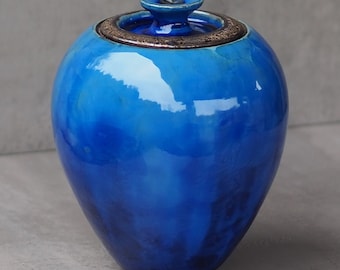 Könnte beinhalten: Eine blaue Keramikvase mit Deckel. Die Vase hat eine glänzende Oberfläche und einen dezenten Marmoreffekt. Der Deckel ist hellbraun mit einem metallischen Schimmer.