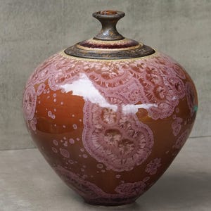 Op de afbeelding: Een ronde keramische pot met een glanzende afwerking, voorzien van een dieproodbruine basiskleur en ingewikkelde, lichtroze kristallijne glazuurpatronen die op bloemen lijken. Het bevat een bijpassend deksel met een donkere, getextureerde knop.
