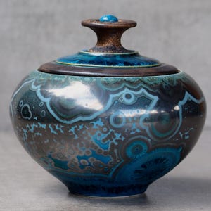Op de afbeelding: Een ronde keramische pot met deksel, voorzien van een glanzende afwerking met complexe, wervelende patronen in tinten diepblauw, taling en donkerbruin. Het deksel heeft een donkere, getextureerde rand en een centrale knop met een blauw accent.