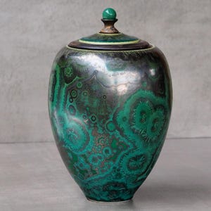 Op de afbeelding: Een keramische pot met deksel voorzien van een levendig groen en zwart glazuur geïnspireerd op malachiet. De pot heeft een afgerond lichaam en een kleine, bolvormige groene knop bovenop het deksel. Het oppervlak vertoont ingewikkelde, wervelende patronen in smaragdgroene en donkere bosgroene tinten, wat zorgt voor een gestructureerd, organisch uiterlijk over de gehele buitenkant.