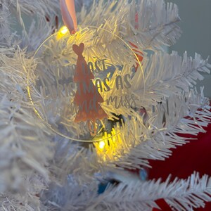 May include: A clear, circular Christmas ornament with the text "Last Christmas as Miss 2023" and a peach-coloured Christmas tree design. The ornament hangs from a pink ribbon on a white Christmas tree with yellow lights.