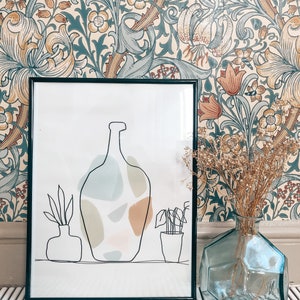 May include: A framed art print with a black border shows a minimalist line drawing of vases and abstract shapes in muted colours. A clear, geometric glass vase holds dried flowers, set against a floral wallpaper background.