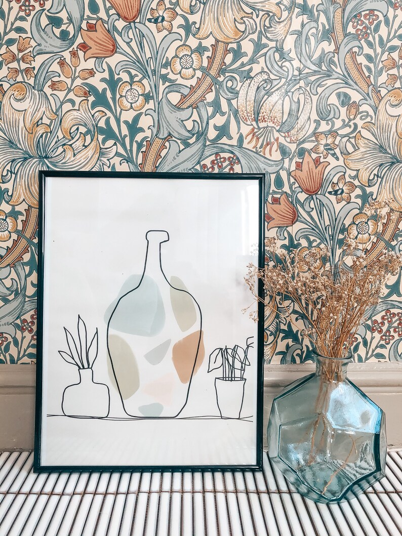 May include: A framed art print with a black border shows a minimalist line drawing of vases and abstract shapes in muted colours. A clear, geometric glass vase holds dried flowers, set against a floral wallpaper background.
