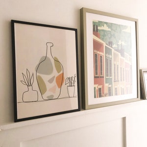 May include: Two framed art prints. One has a black line drawing of a vase with abstract shapes in muted colours. The other depicts a row of colourful houses with a green and white patterned sky.
