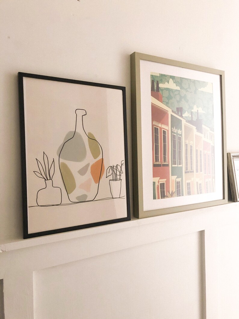 May include: Two framed art prints. One has a black line drawing of a vase with abstract shapes in muted colours. The other depicts a row of colourful houses with a green and white patterned sky.