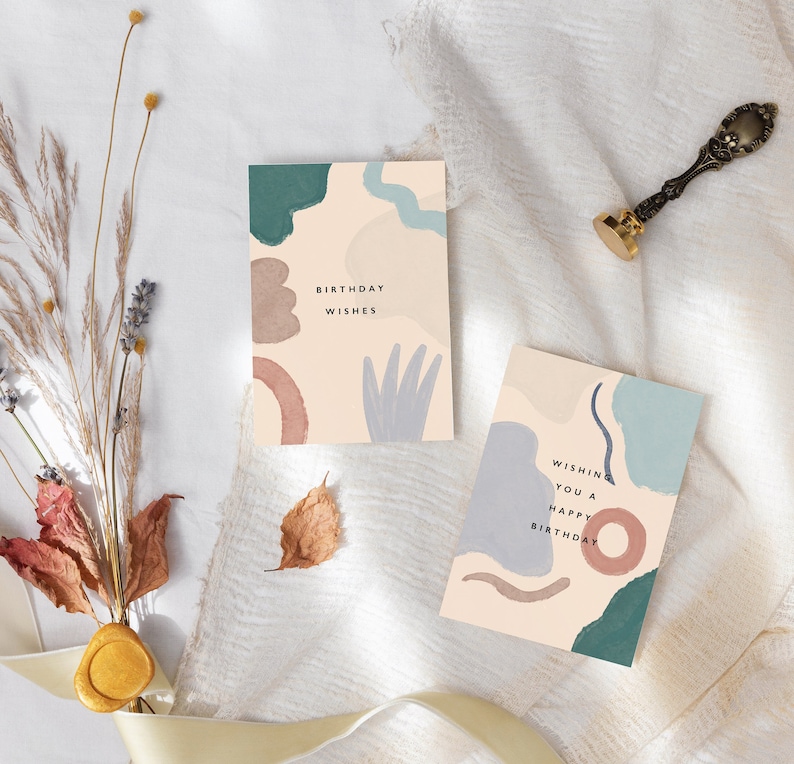 May include: Two birthday cards with abstract watercolour designs in muted tones of teal, brown, and grey. One card reads "Birthday Wishes," the other says "Wishing you a Happy Birthday." A decorative wax seal and dried flowers are also present.