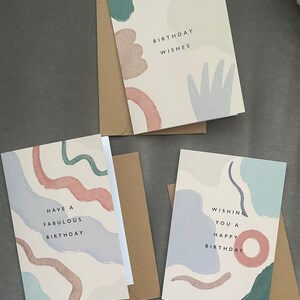 May include: Three birthday cards with abstract watercolour designs in muted tones of blue, green, and pink. Each card has a different message: "Birthday Wishes", "Have a Fabulous Birthday", and "Wishing You a Happy Birthday". Kraft paper envelopes accompany each card.