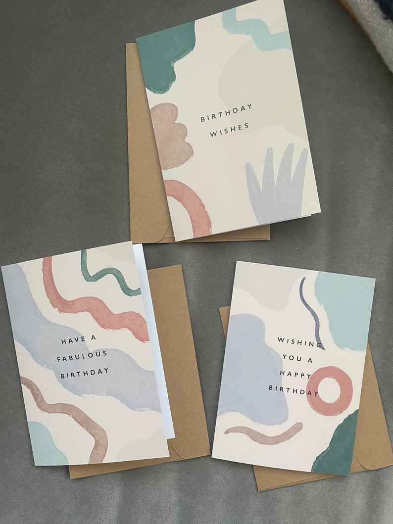 May include: Three birthday cards with abstract watercolour designs in muted tones of blue, green, and pink. Each card has a different message: "Birthday Wishes", "Have a Fabulous Birthday", and "Wishing You a Happy Birthday". Kraft paper envelopes accompany each card.