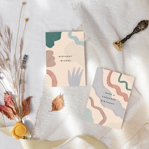 May include: Two birthday cards with abstract watercolour designs in teal, brown, and blue on a cream background. One card reads "Birthday Wishes," the other says "Have a Fabulous Birthday." A gold wax seal and dried flowers are also present.