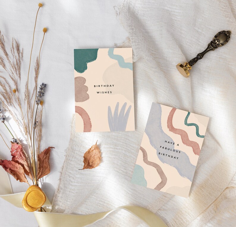 May include: Two birthday cards with abstract watercolour designs in teal, brown, and blue on a cream background. One card reads "Birthday Wishes," the other says "Have a Fabulous Birthday." A gold wax seal and dried flowers are also present.