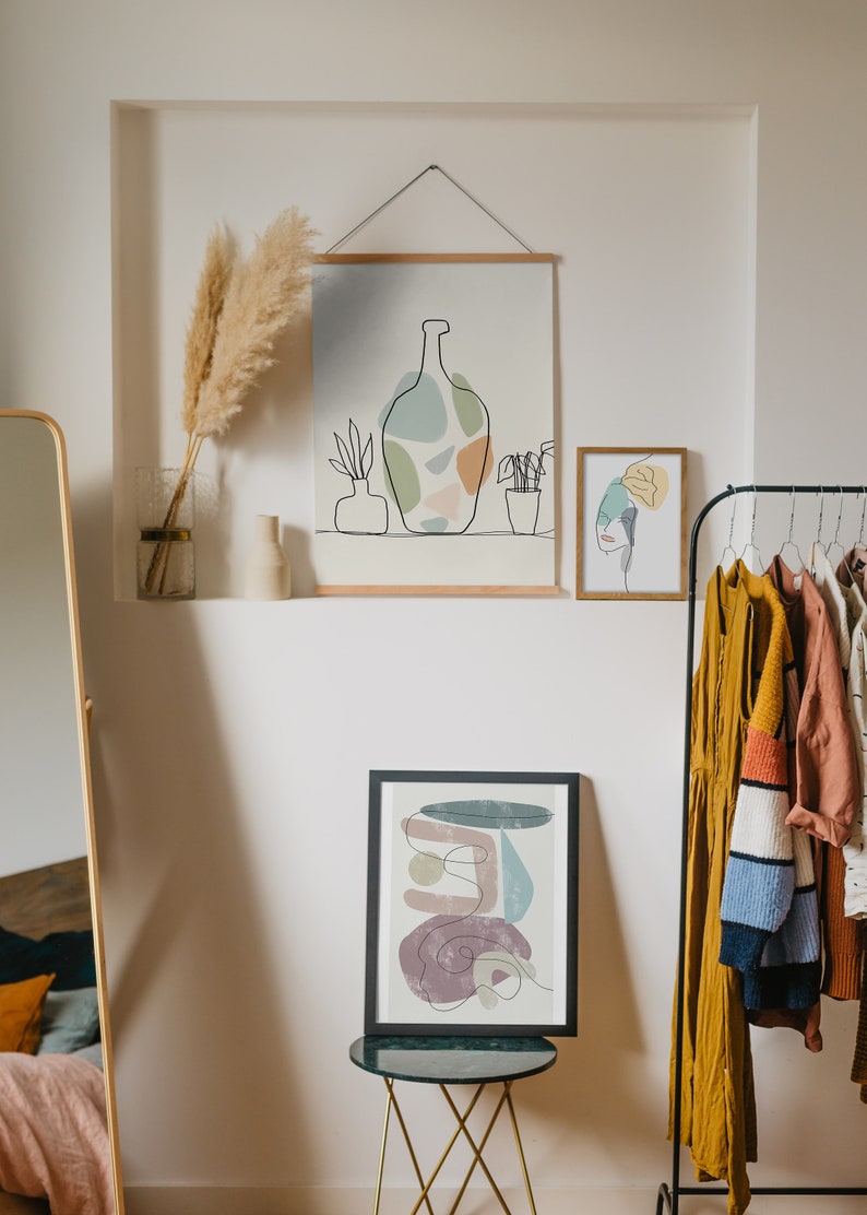 May include: A room interior with framed abstract art prints and a clothes rail. The art includes line drawings of a vase, plants, and faces, with a muted colour palette. A full-length mirror and decorative pampas grass in a vase are also visible.